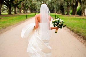 A bride in white walking down the street with her back turned, holding flowers