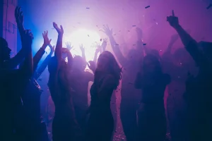 People dancing at a party with raised hands and confetti under purple lighting.